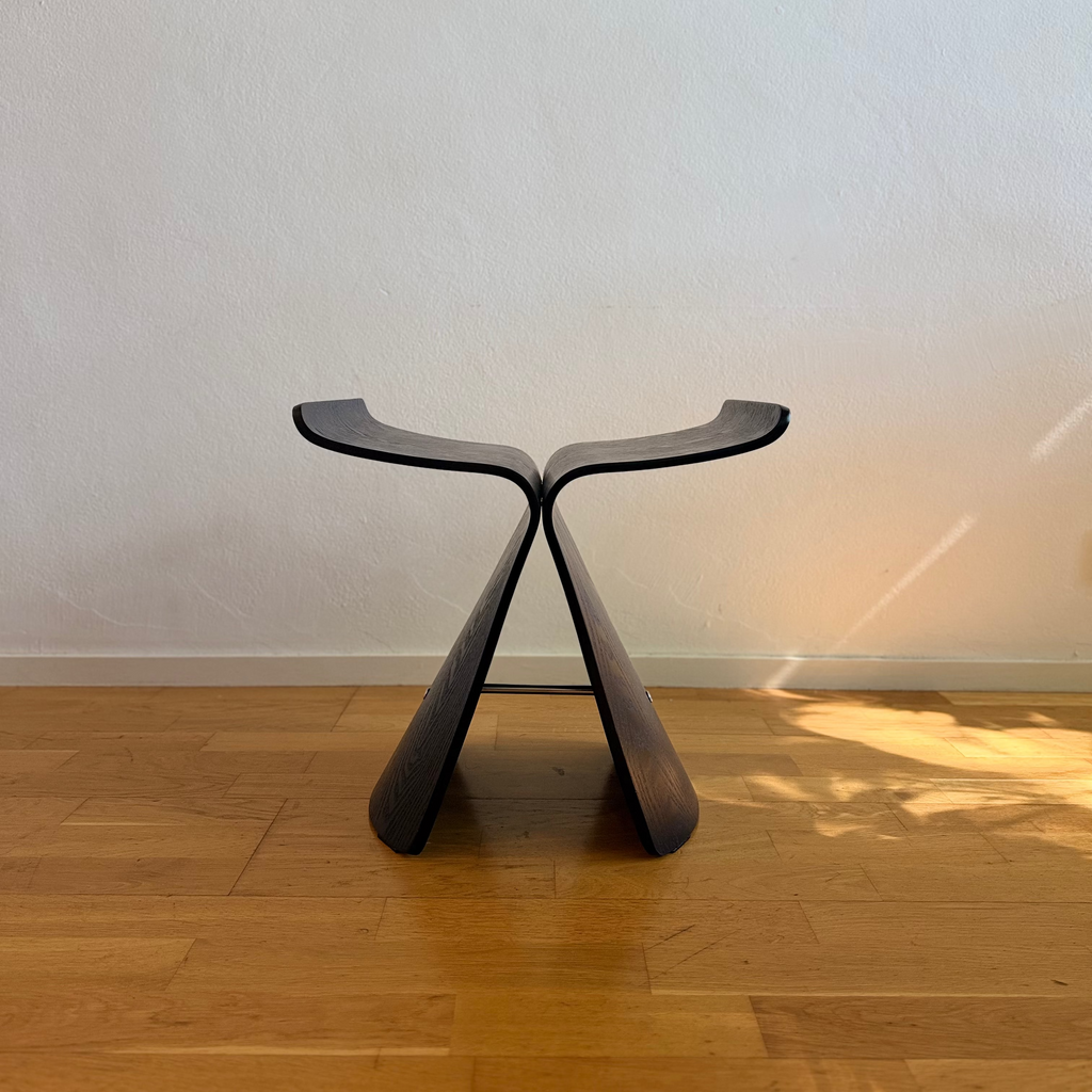 A modern sculptural wooden stool with a dark finish, standing on a light wood floor against a textured off-white wall.