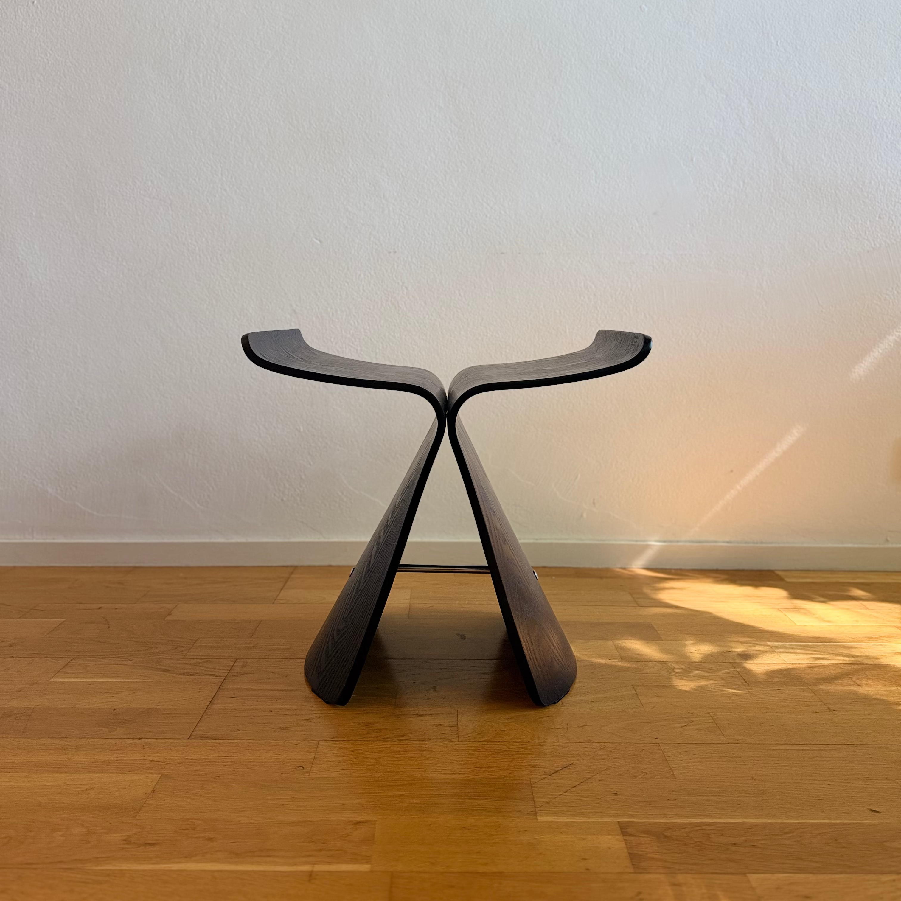 A modern sculptural wooden stool with a dark finish, standing on a light wood floor against a textured off-white wall.