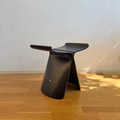 A modern sculptural wooden stool with a curved seat and backrest, shown on a light wood floor against a plain off-white wall.