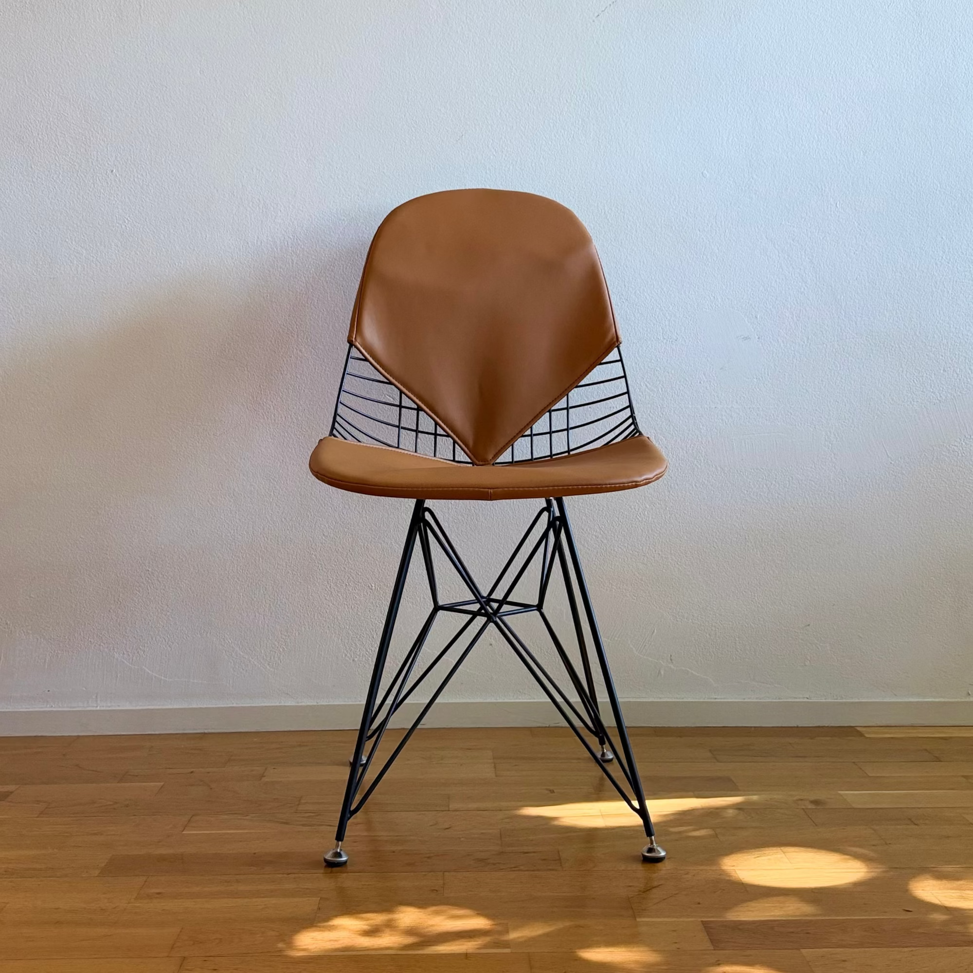 A mid-century modern style chair with a brown leather seat and backrest, supported by a black wire frame and splayed legs, set on a light wood floor against a plain off-white wall.