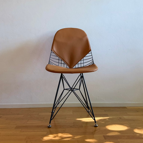 A mid-century modern style chair with a brown leather seat and backrest, supported by a black wire frame and splayed legs, set on a light wood floor against a plain off-white wall.