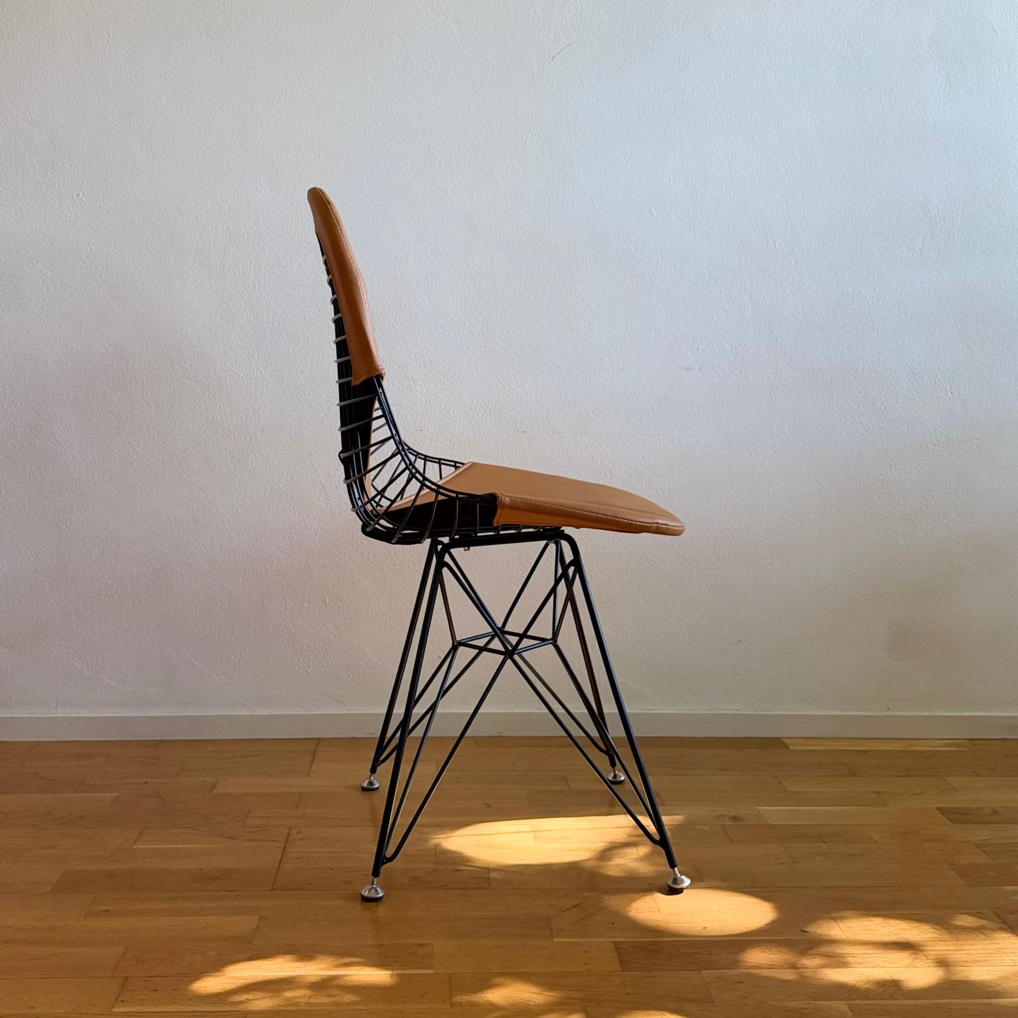 A mid-century modern wire chair with a light brown leather seat and backrest, set on a light wood floor against a plain off-white wall with natural leaf shadows.