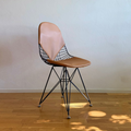 A mid-century modern wireframe chair with a light brown leather seat and backrest, supported by a black metal wire frame and chrome legs, set against a textured off-white wall and light wood floor.