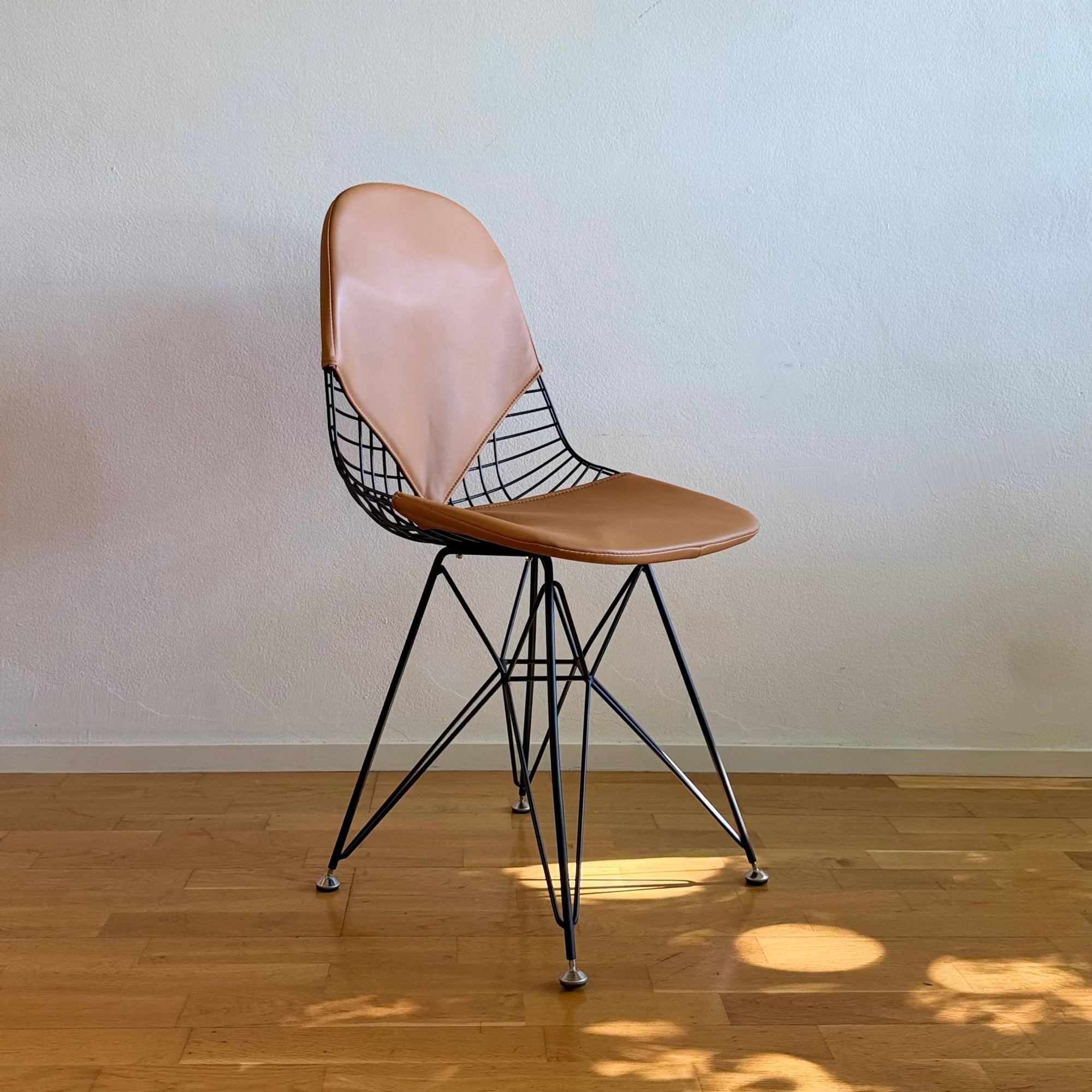 A mid-century modern wireframe chair with a light brown leather seat and backrest, supported by a black metal wire frame and chrome legs, set against a textured off-white wall and light wood floor.