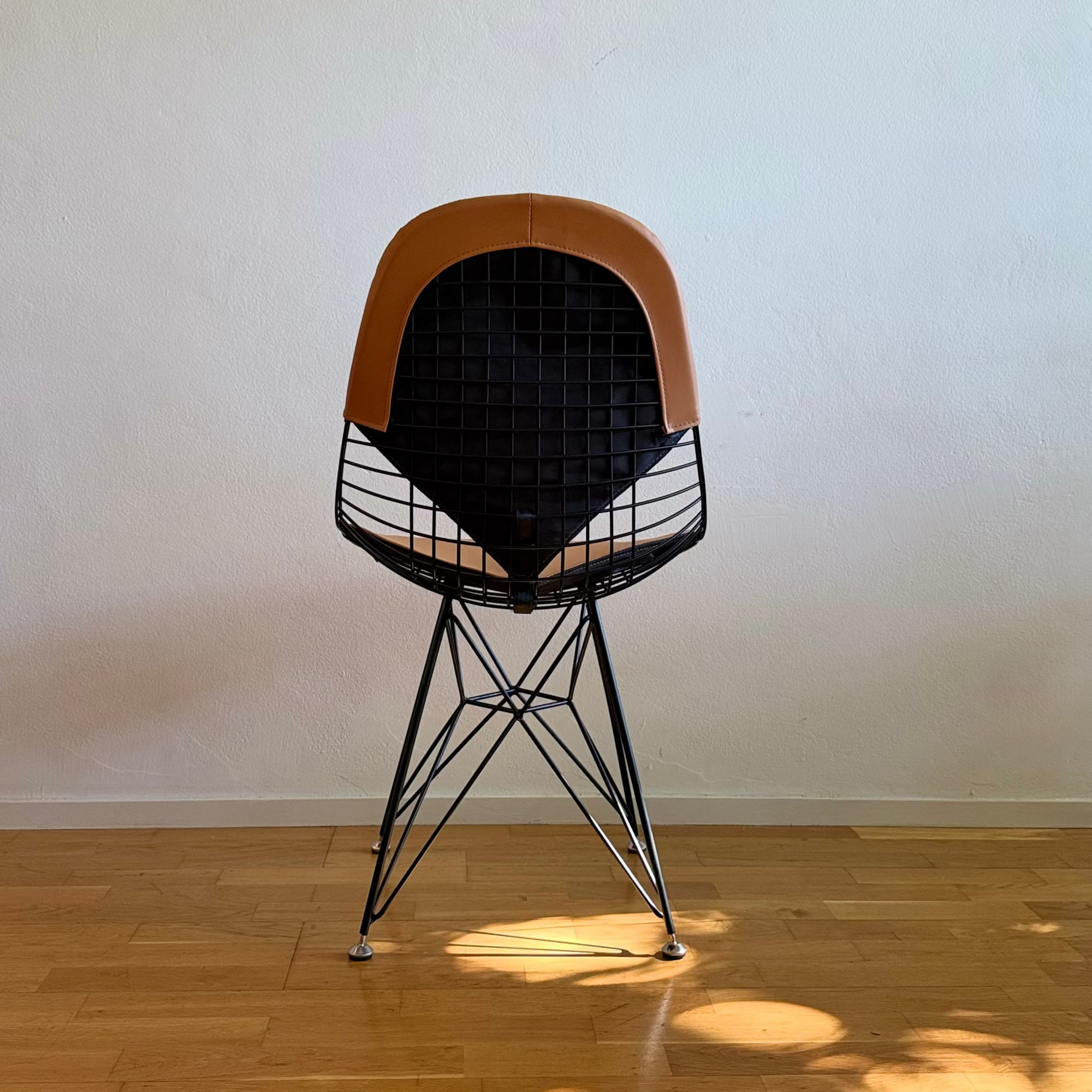 A mid-century modern wire chair with a light brown leather seat and backrest, set on a light wood floor against a plain off-white wall with natural leaf shadows.