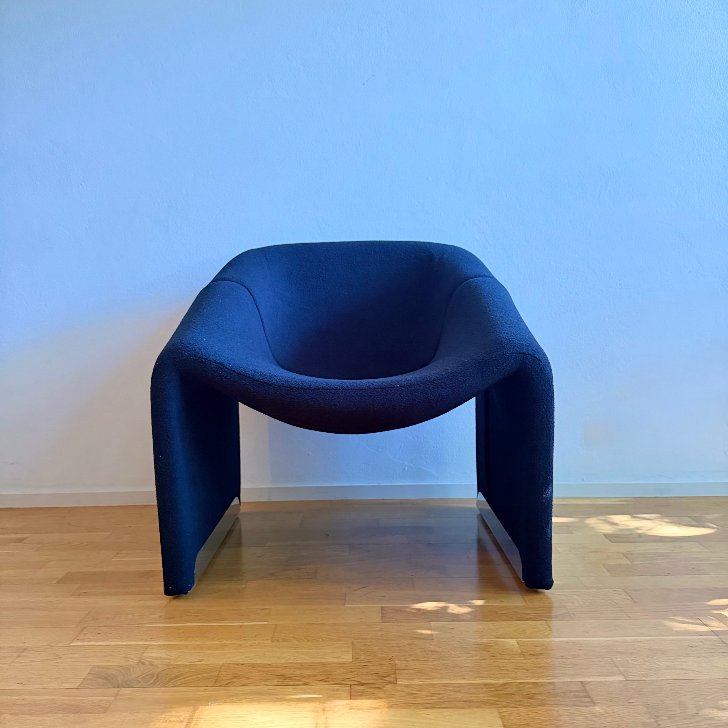A modern sculptural armchair upholstered in black fabric, set against a light blue wall on a light wood floor.