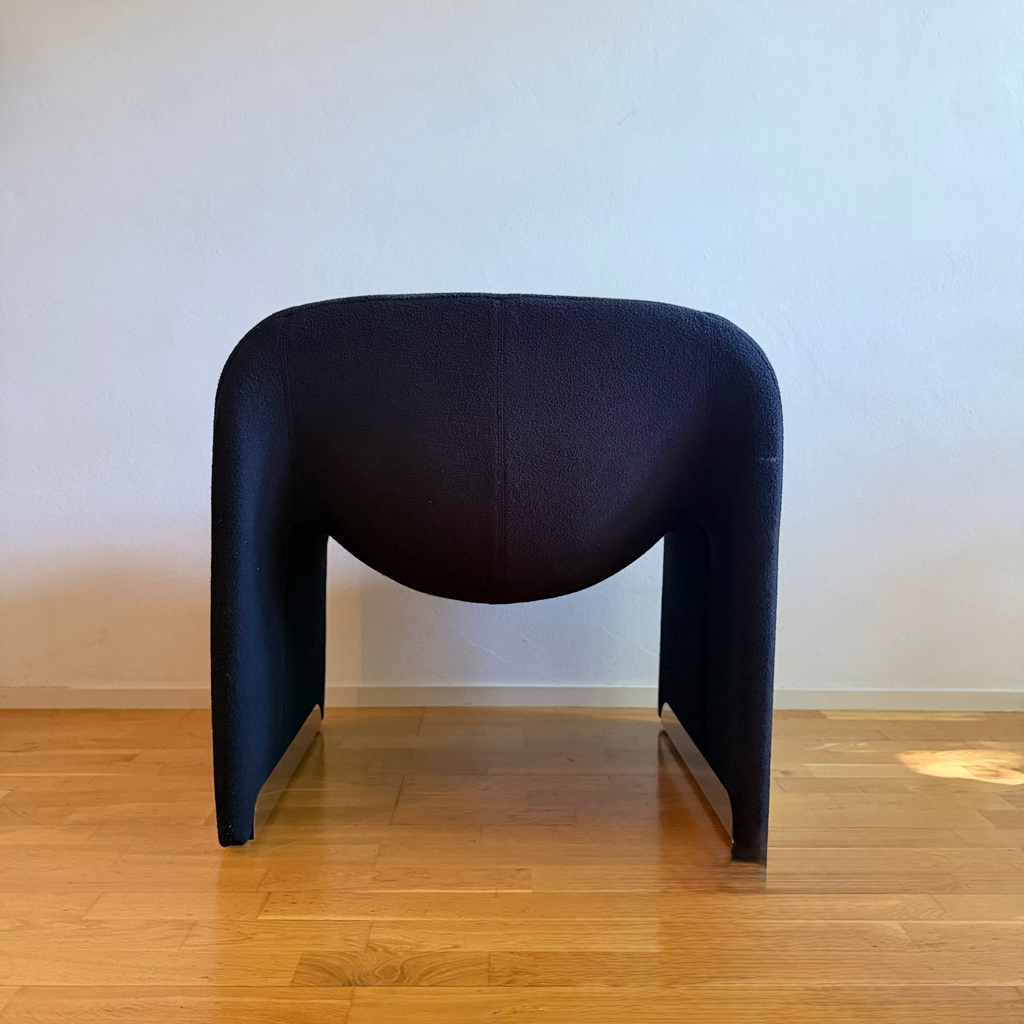 A modern sculptural armless chair upholstered in a dark navy blue textured fabric, set against a plain off-white wall on a light wood floor.