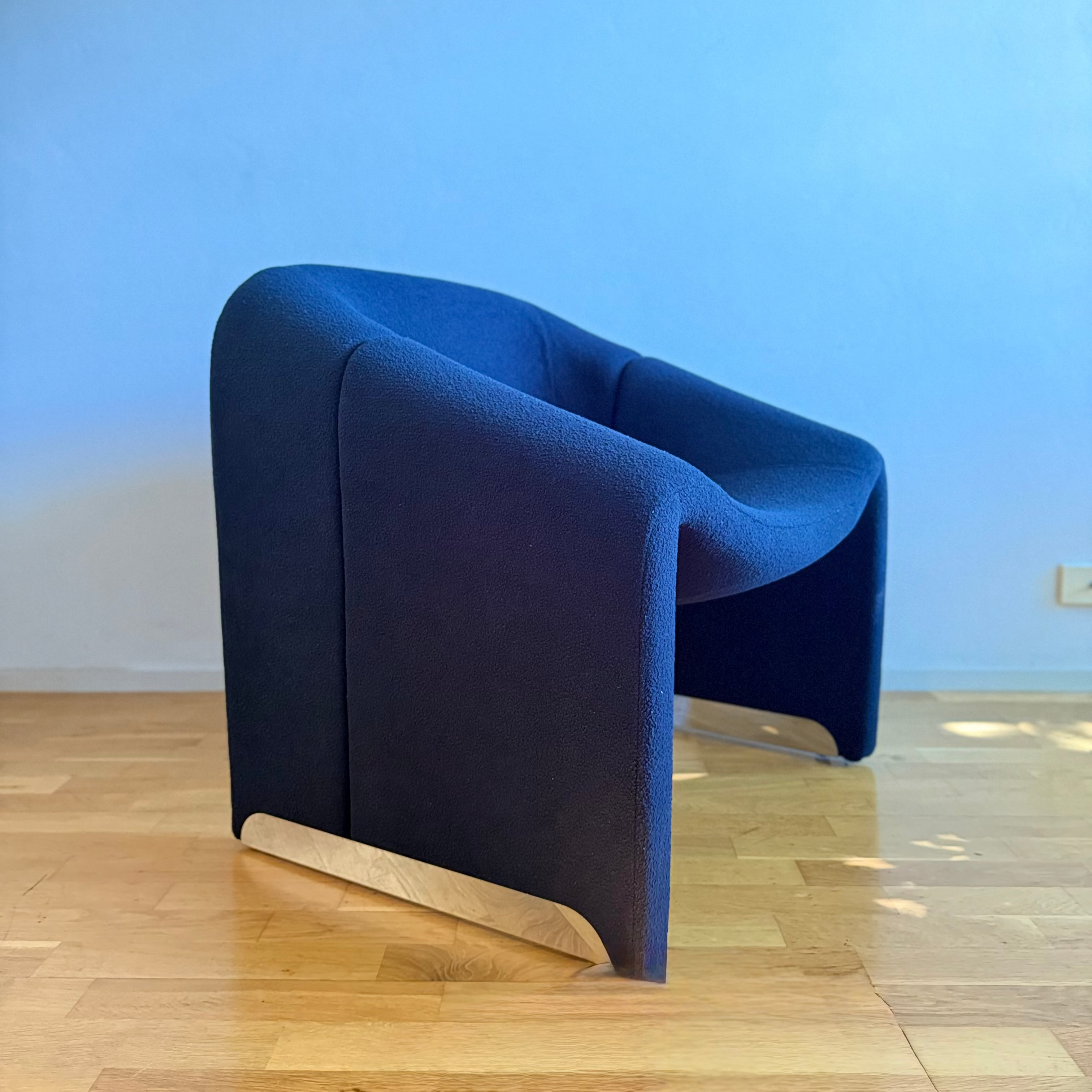 A modern curved armchair upholstered in deep navy blue fabric, featuring a light natural wood base, set on a light wood parquet floor against a solid blue wall.
