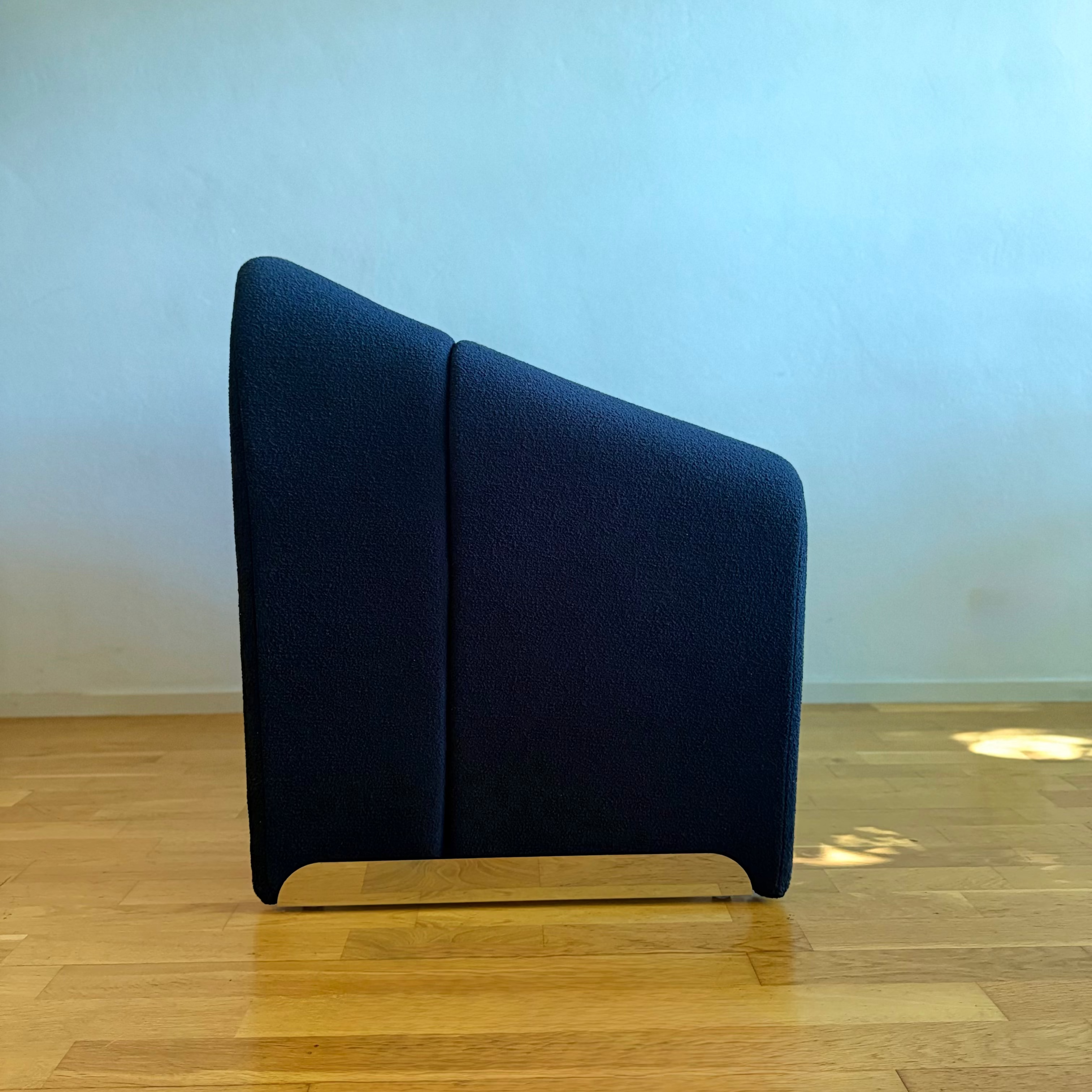 A modern dark blue upholstered accent chair with a slanted backrest and a light wood base, shown on a light wood floor against a pale blue wall.