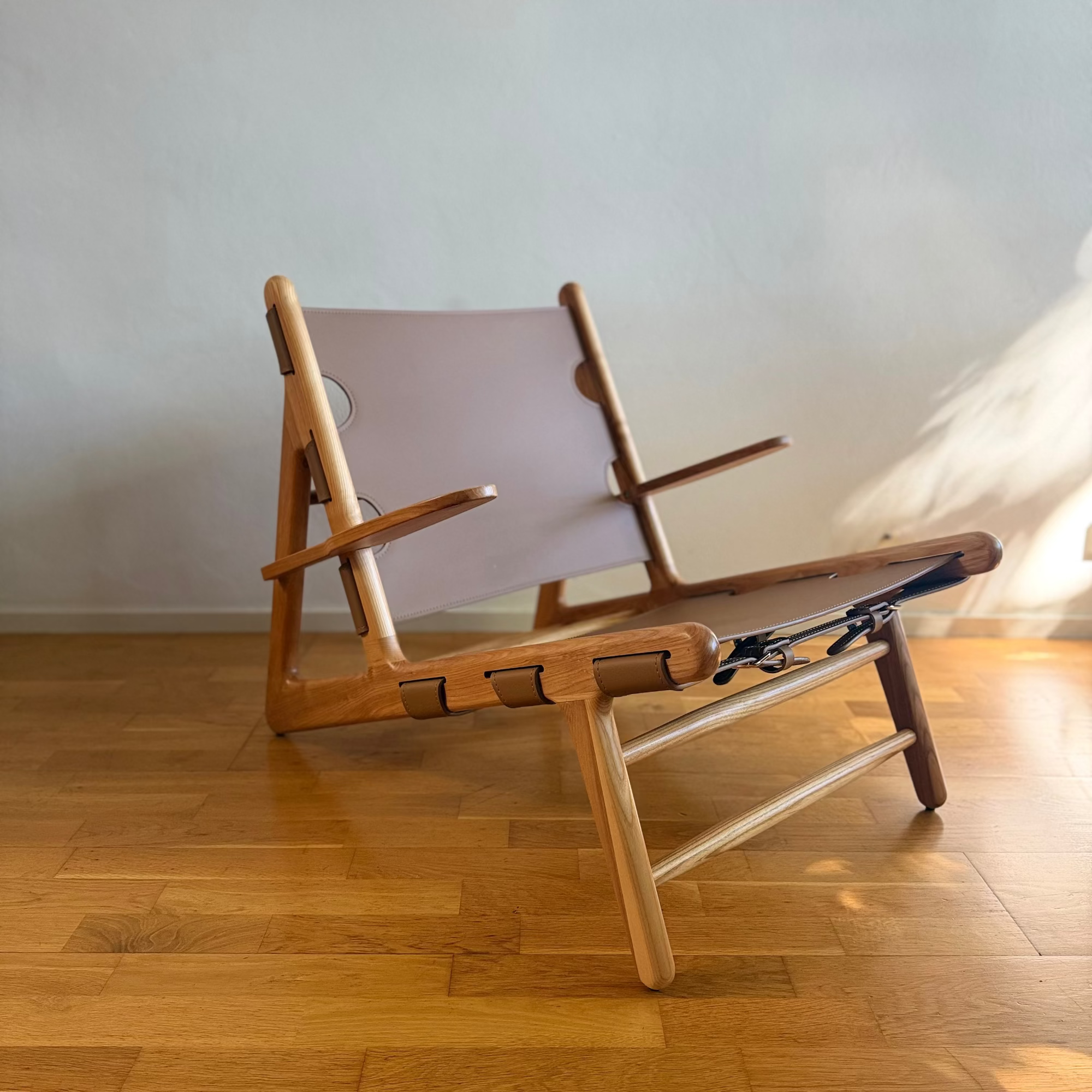 A mid-century modern wooden lounge chair with a light beige canvas sling seat, positioned on a polished wooden parquet floor against a neutral light gray wall.