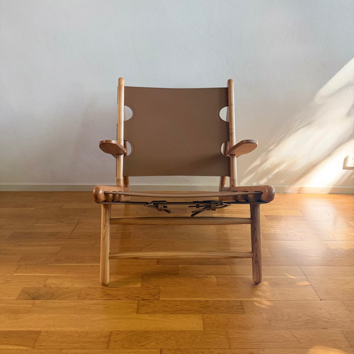 A mid-century modern wooden lounge chair with a brown canvas sling seat and backrest, set on a polished wooden parquet floor against a plain light gray wall.