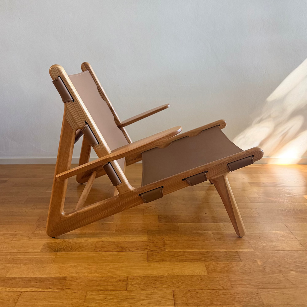 A mid-century modern wooden lounge chair with brown canvas fabric, positioned on a light wood parquet floor against a textured off-white wall with warm sunlight casting shadows.