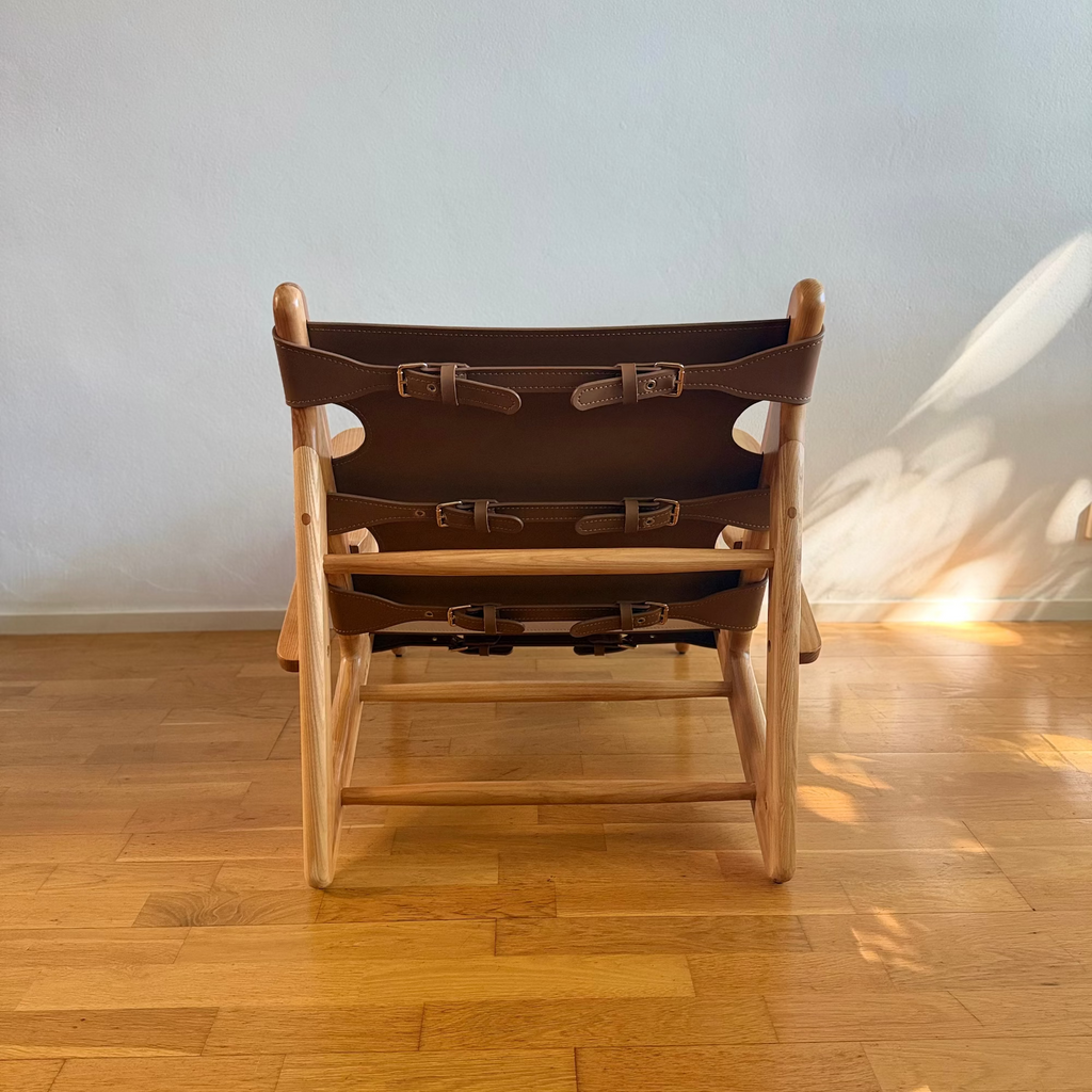 A mid-century modern wooden lounge chair with brown leather straps, photographed from the back in a room with a light gray wall and a warm wood parquet floor.