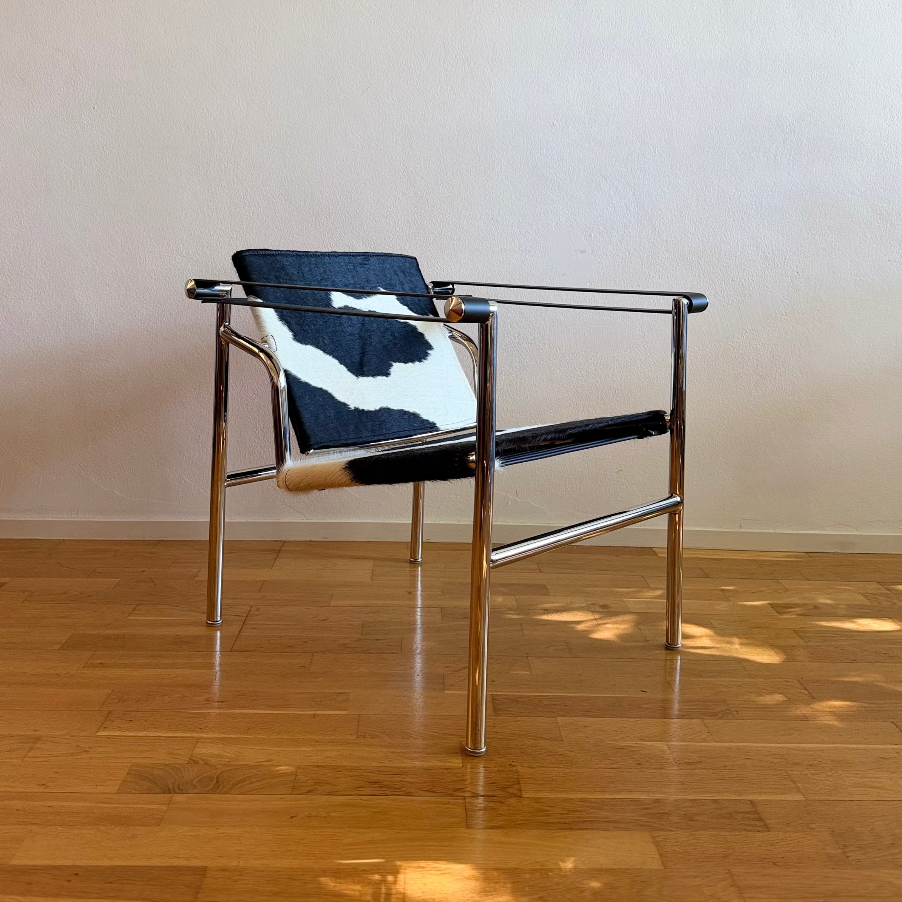 A mid-century modern metal armchair with a polished chrome frame and a black-and-white cowhide seat and backrest, set on a light wood parquet floor against a plain beige wall.