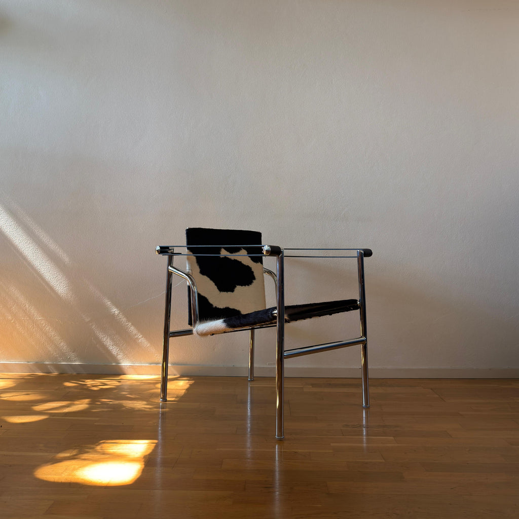 A modern metal-framed chair with a black and white cowhide seat and backrest, set against a plain beige wall on a wooden floor with sunlight casting shadows.