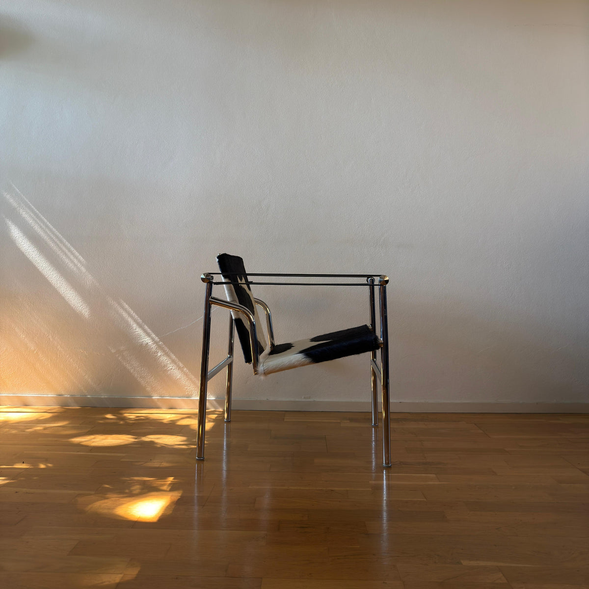 A modern metal chair with black and white patterned seat and back cushions, set against a plain beige wall on a wooden floor with sunlight casting shadows.