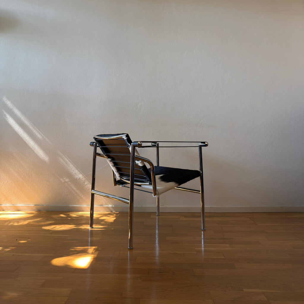 A modern metal-framed chair with black and white woven sling seats, placed on a polished wooden floor against a plain beige wall with warm sunlight and shadows.