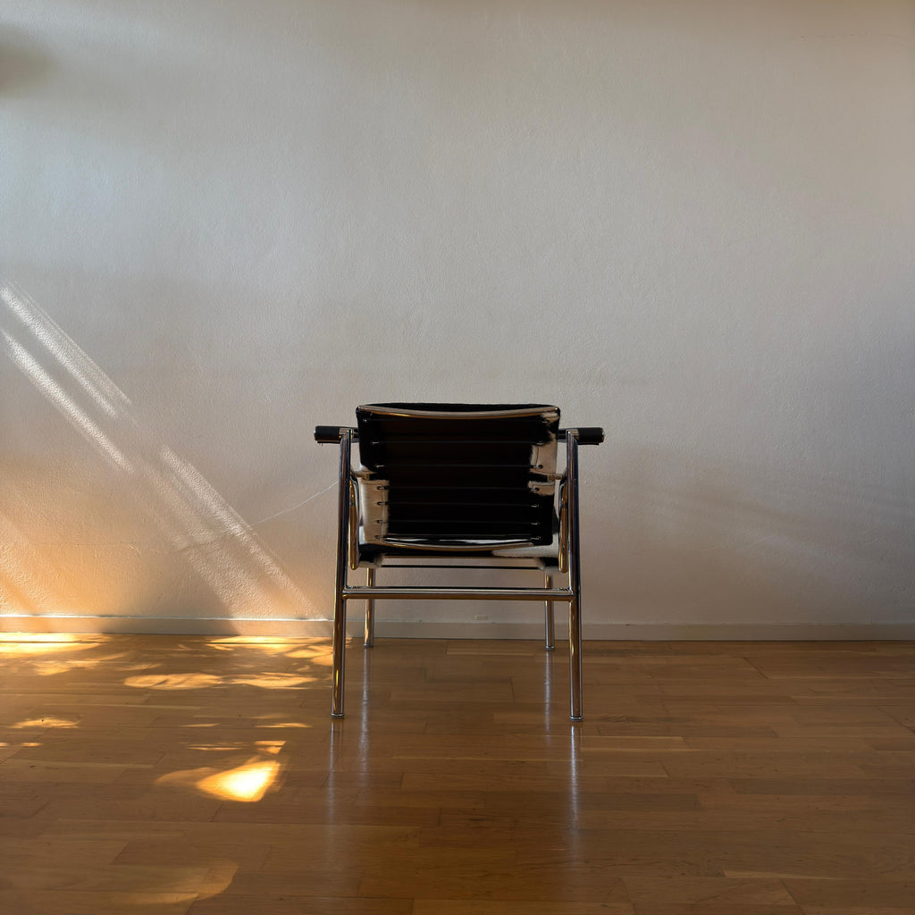 A modern black and silver metal chair is positioned against a plain beige wall on a wooden floor, with warm sunlight casting diagonal shadows on the wall and floor.