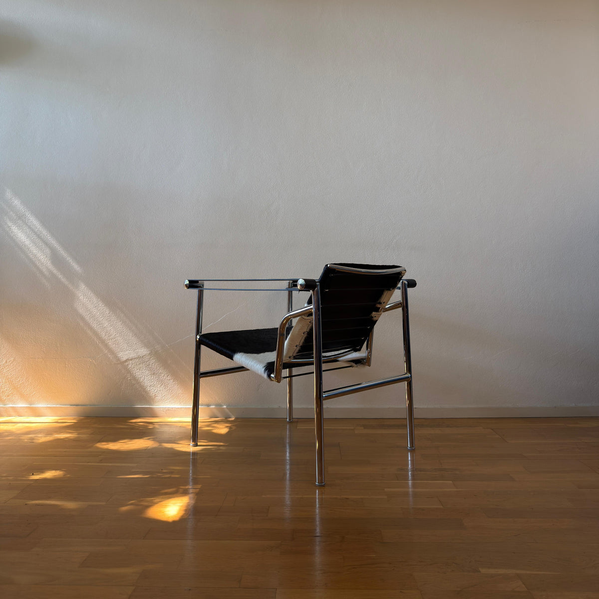 A modern armchair with a black woven sling seat and back, supported by a chrome metal frame, set against a plain beige wall on a polished wooden floor with sunlight casting shadows.