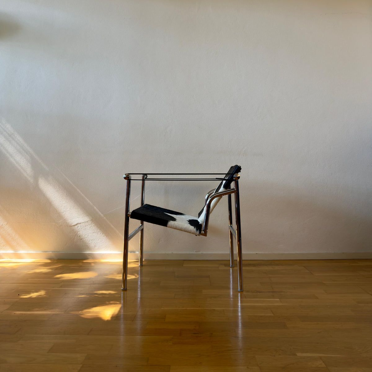 A modern metal chair with a black and white patterned seat cushion, positioned in a minimalist room with a beige wall, wooden floor, and warm sunlight streaming in from the side.