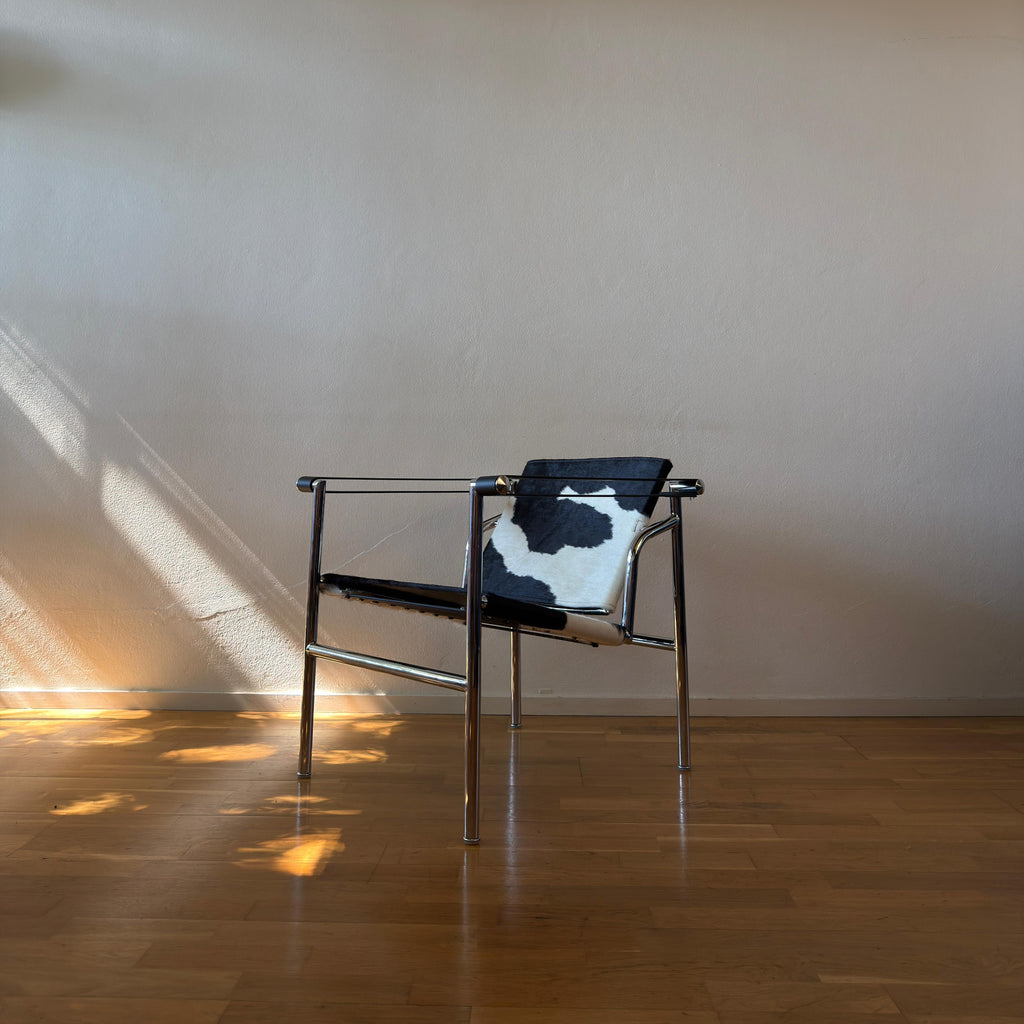 A modern metal-framed armchair with a black and white cowhide seat and backrest, set against a plain beige wall on a polished wooden floor with sunlight casting shadows.
