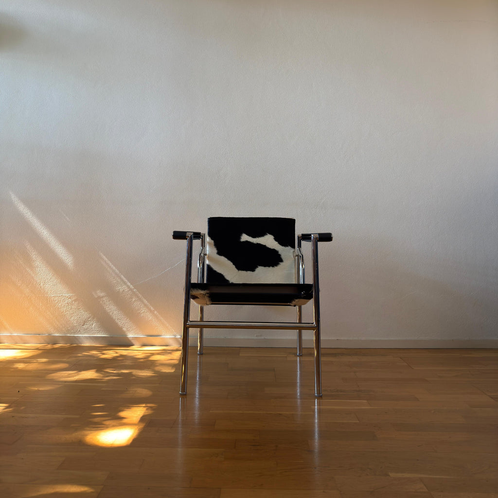 A mid-century modern chair with a black and white cowhide seat cushion and chrome metal frame, set against a plain beige wall on a wooden floor with sunlight casting shadows.