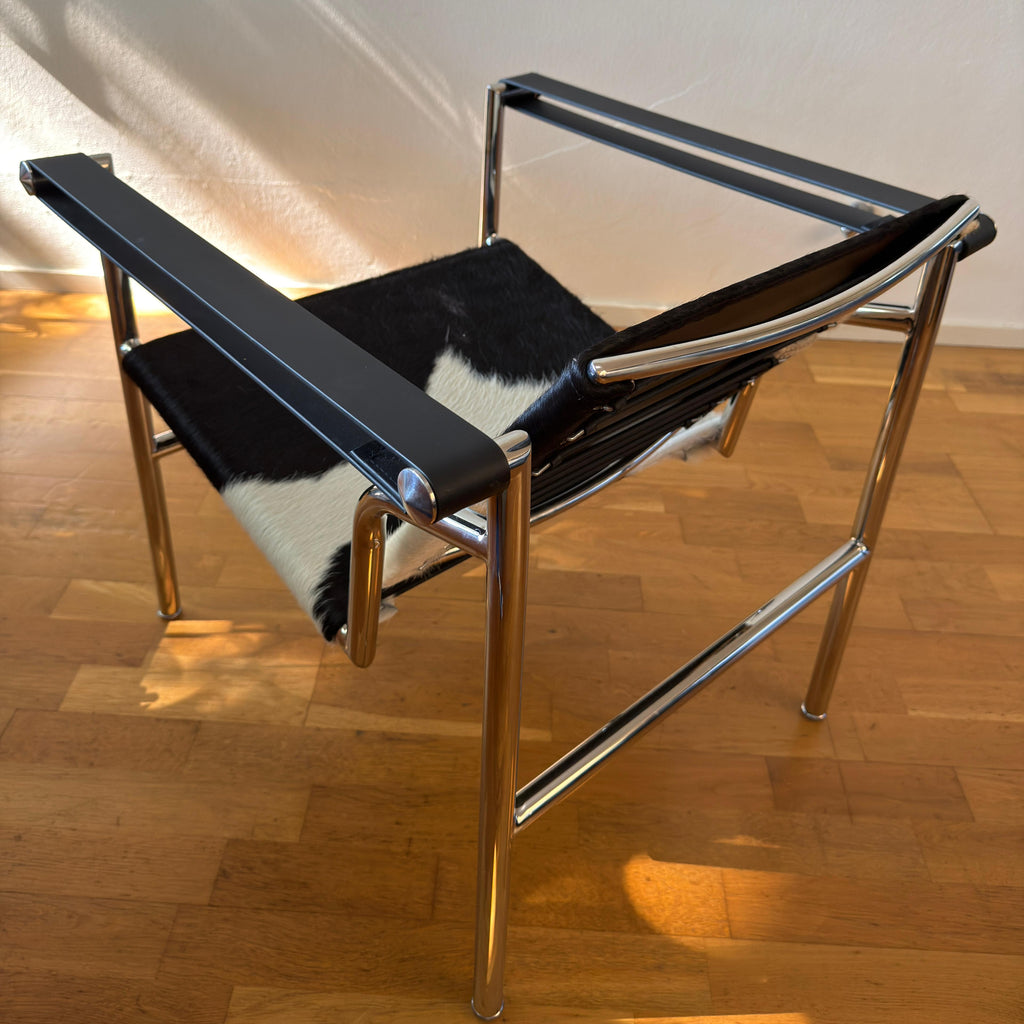 A modern armchair with a polished chrome tubular steel frame, black leather armrests and backrest, and a black-and-white cowhide seat, shown on a wooden parquet floor against a white wall.