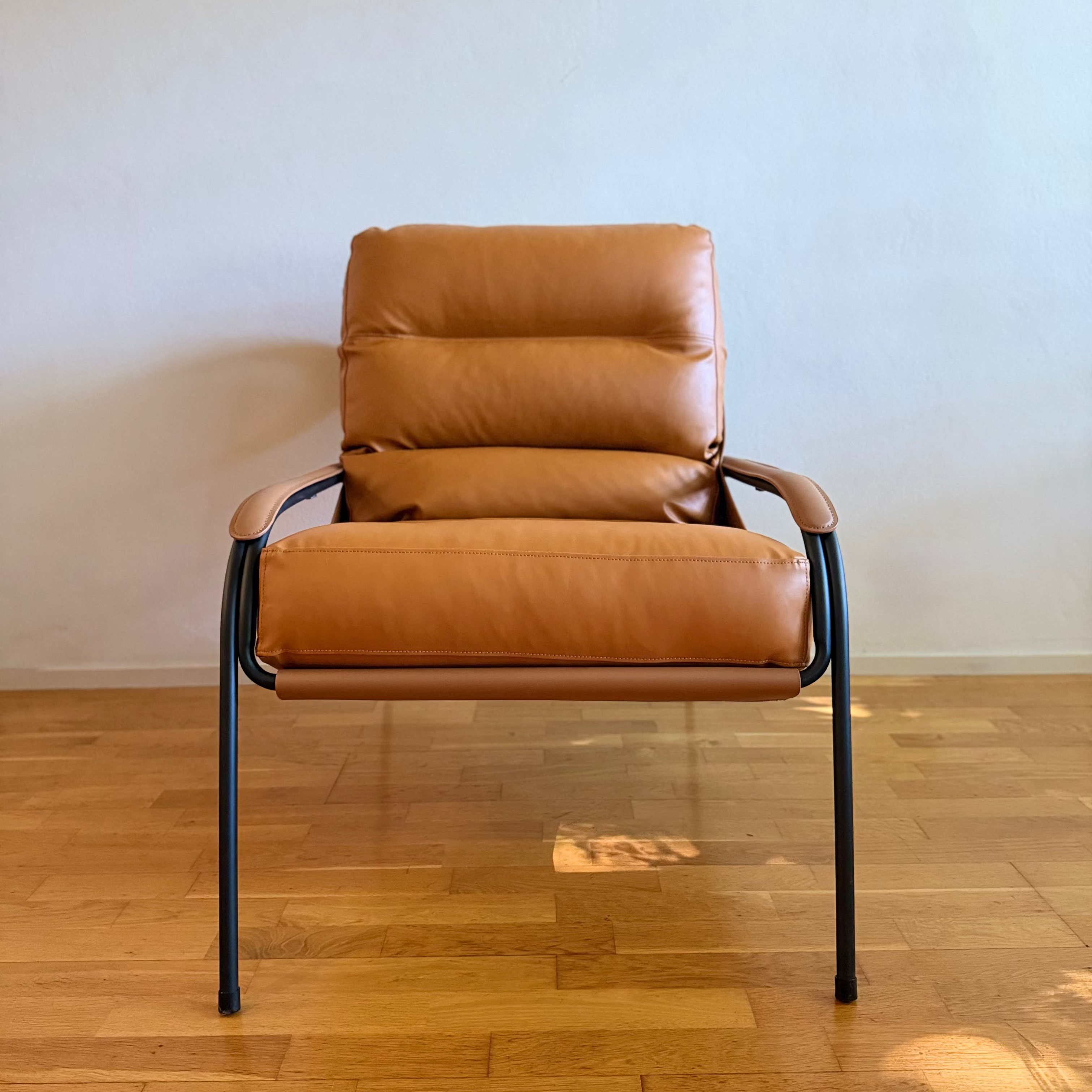 A mid-century modern accent chair with tan leather cushions and a black metal frame, set against a plain light gray wall on a wooden floor.