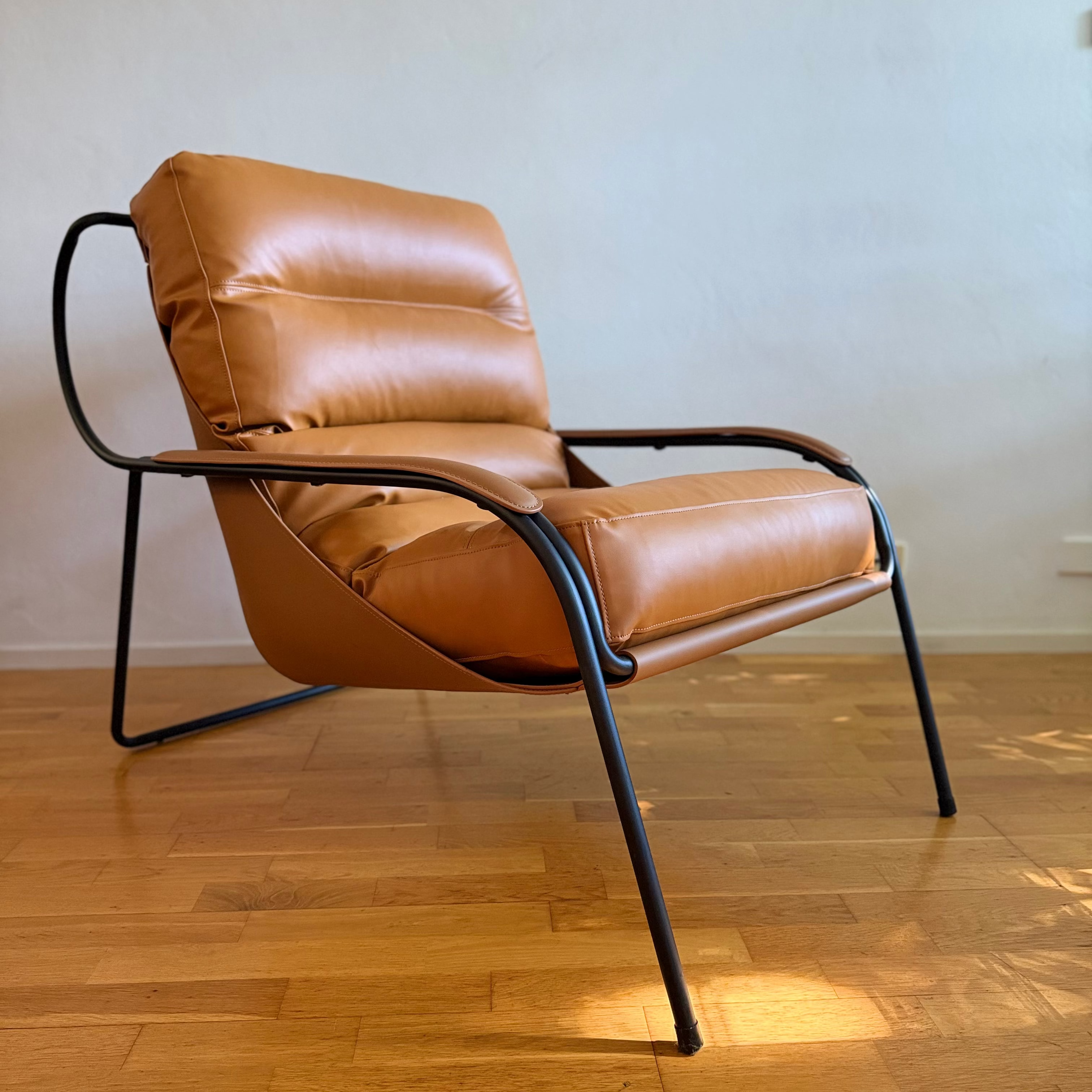 A mid-century modern accent chair with tan leather cushions and a black metal frame, set on a wooden floor against a plain wall.