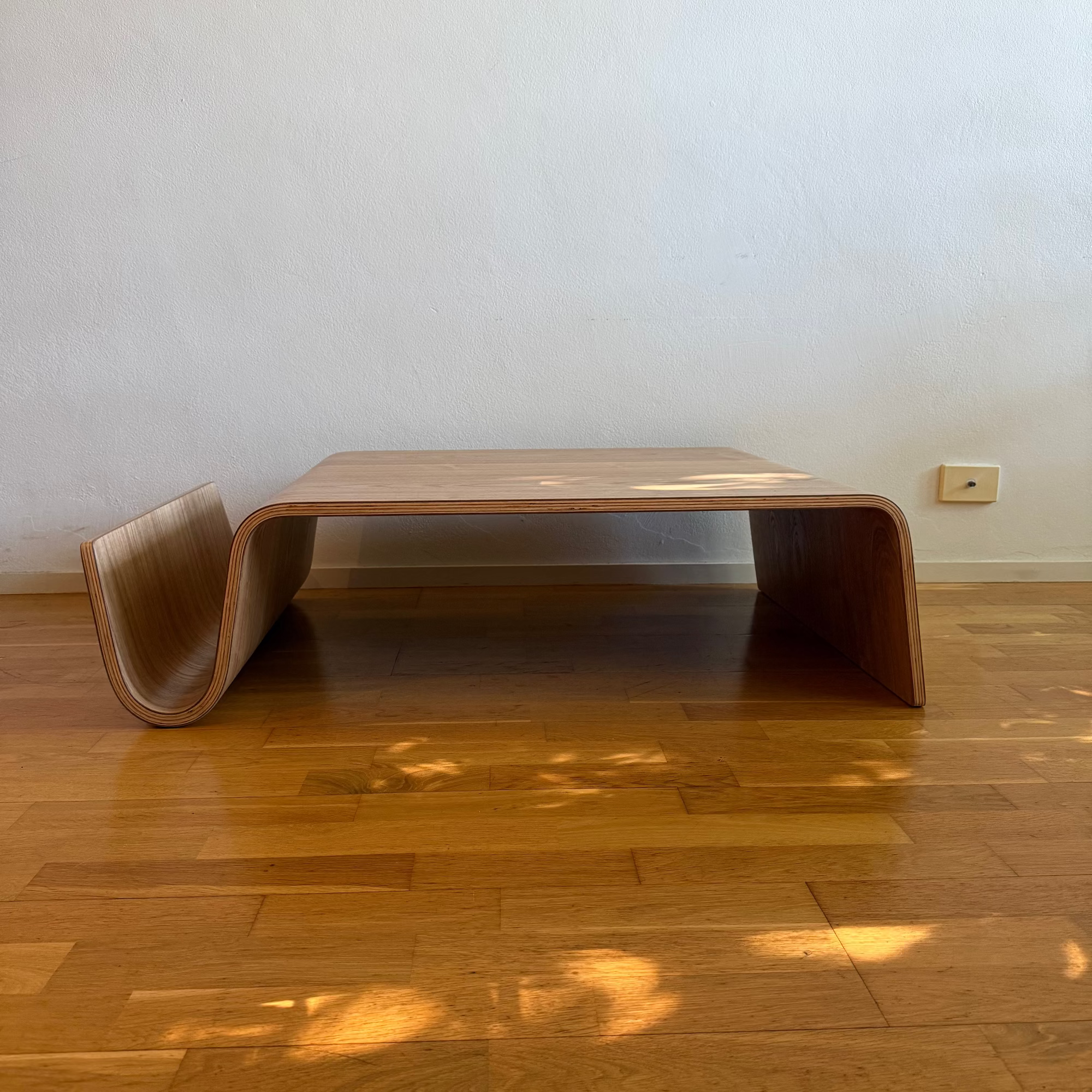 A low wooden table with a curved, shell-like structure and open center, placed on a polished hardwood floor against a plain off-white wall.