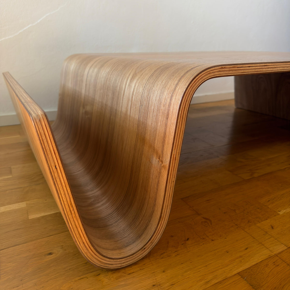 A modern wooden coffee table with a sculptural, curved shell design made from layered plywood, shown on a wooden floor against a plain wall.