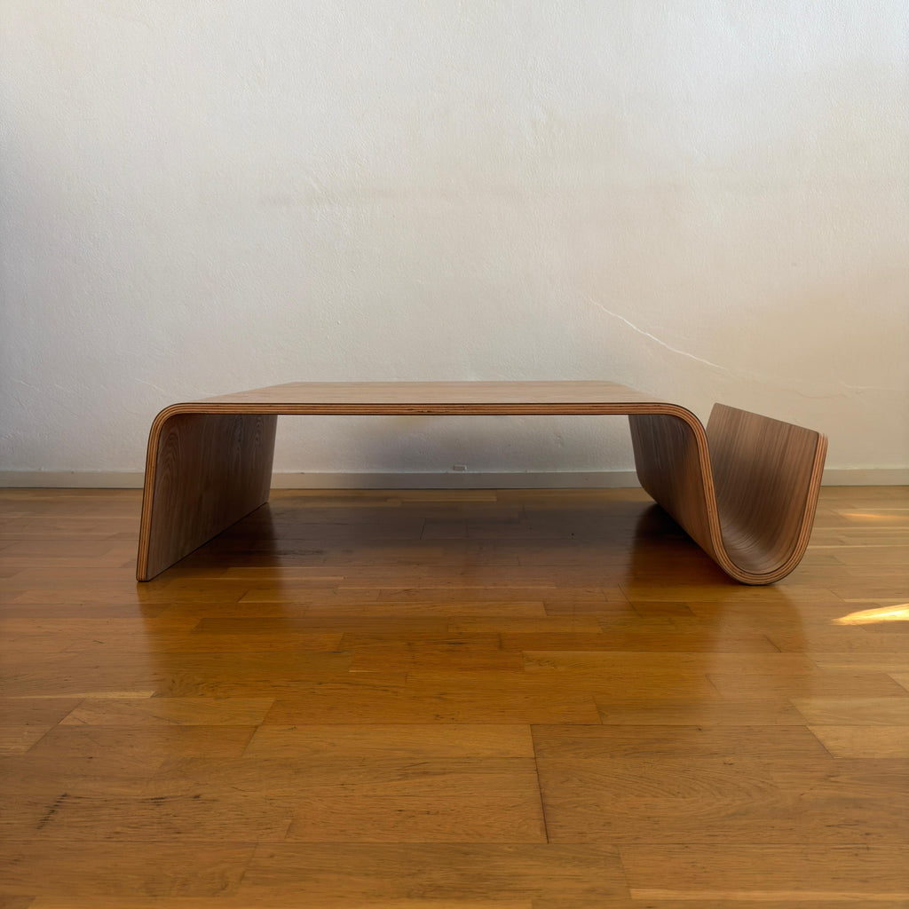 A modern low wooden coffee table with a curved, shell-like design and a warm medium-brown finish, set on a polished wood floor against a plain off-white wall.