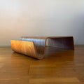 A modern curved wooden bench with a sculptural, wave-like design, positioned on a polished wooden floor against a plain beige wall.