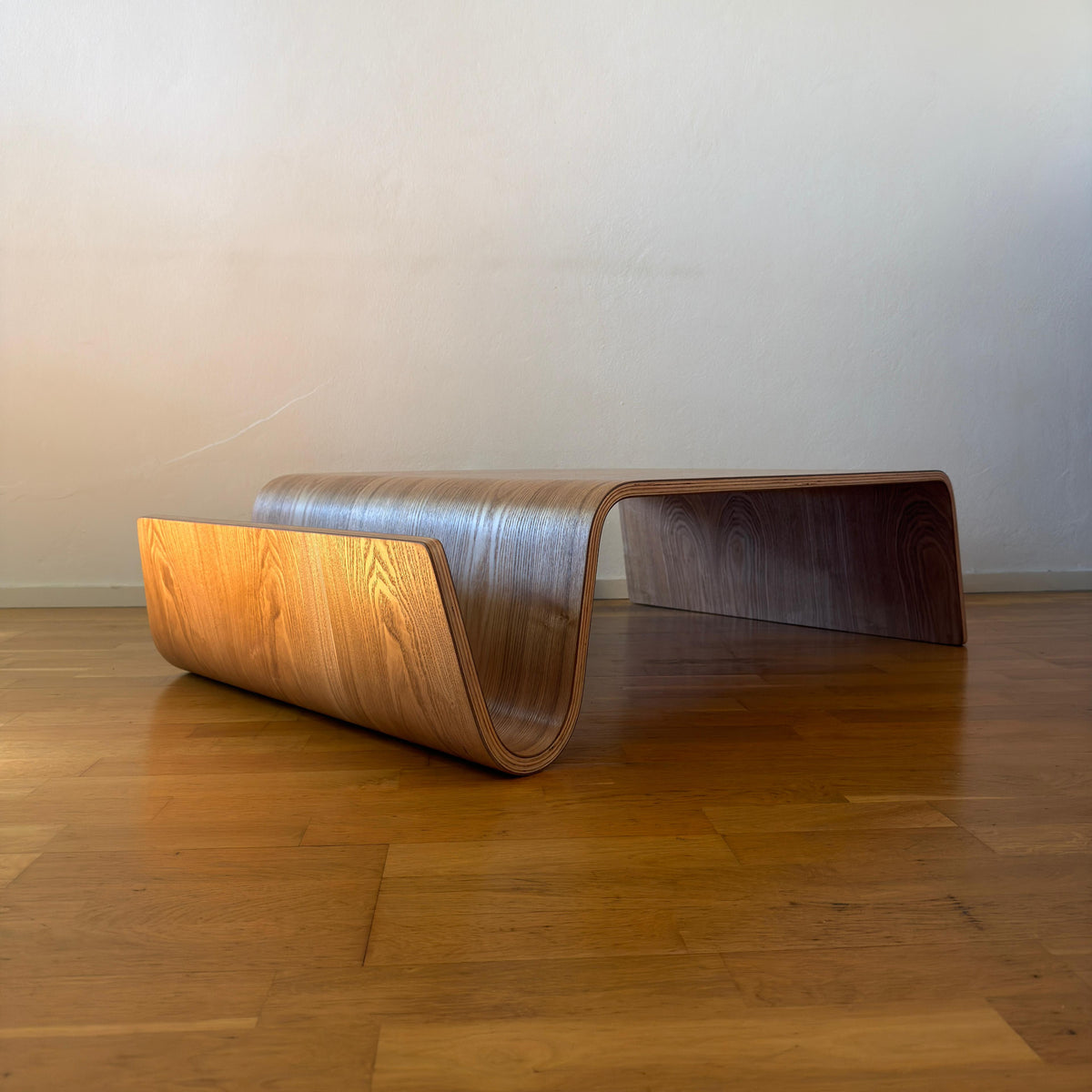 A modern curved wooden bench with a sculptural, wave-like design, positioned on a polished wooden floor against a plain beige wall.