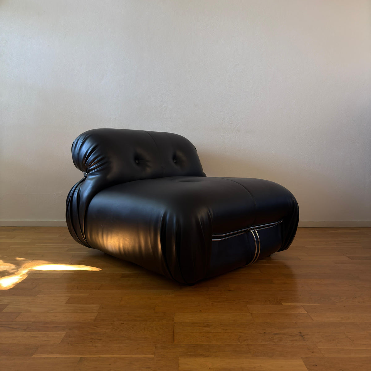 A black leather chaise lounge chair with a tufted backrest, placed on a polished wooden floor against a plain beige wall.