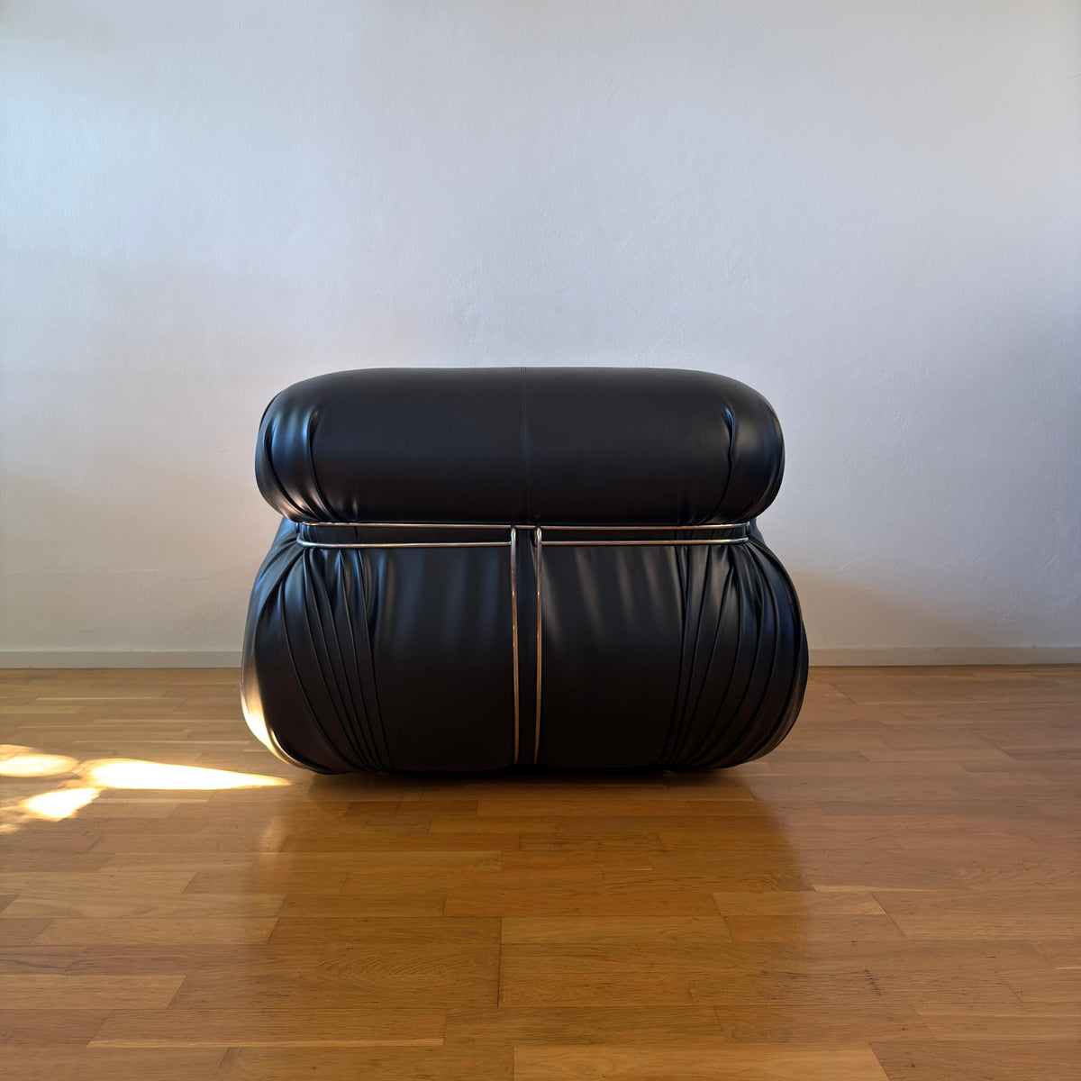A modern black leather ottoman or small stool with a rounded rectangular top and gathered, ruched sides, shown on a polished wood floor against a plain light wall.