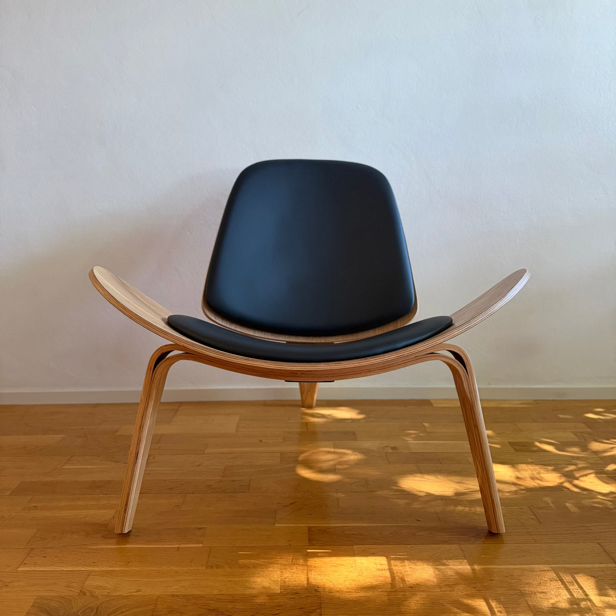 A classic mid-century modern lounge chair with a curved light wood shell and black vinyl seat and backrest, set on a wooden floor against a plain wall.