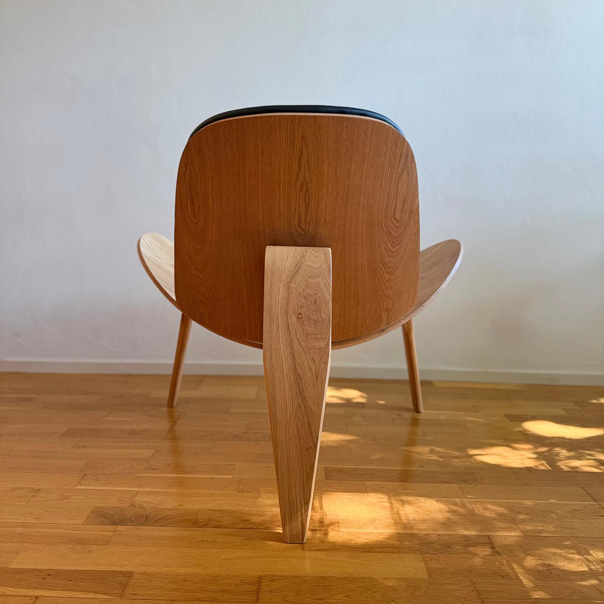 A mid-century modern wooden lounge chair with a curved backrest and sculptural tapered legs, shown from the back in a room with light wood floors and a plain wall.