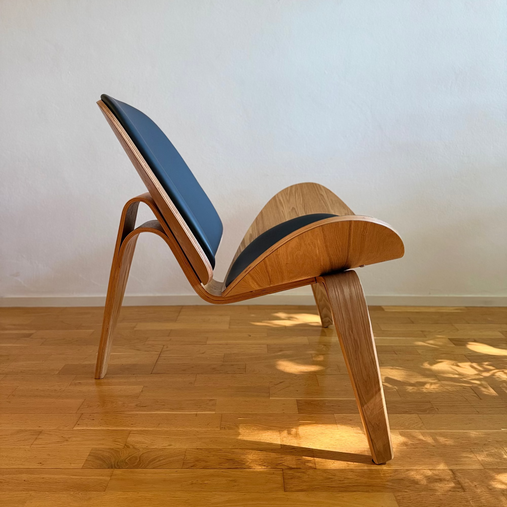 A mid-century modern wooden lounge chair with blue and black seat cushions, positioned on a light wood parquet floor against a plain off-white wall.